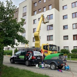 Szuru-Buru Robert Suchodolski - Pracownik na podnośniku koszowym czyści elewację budynku wielorodzinnego; widoczny samochód firmowy z logo 'Szuru Buru' i przyczepka ze sprzętem do czyszczenia.