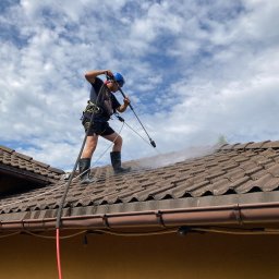 Szuru-Buru Robert Suchodolski - Pracownik w kasku i uprzęży, stojący na brązowym, ceramicznym dachu i spryskujący go wodą pod ciśnieniem z lancy, z widoczną parą wodną w słoneczny dzień.