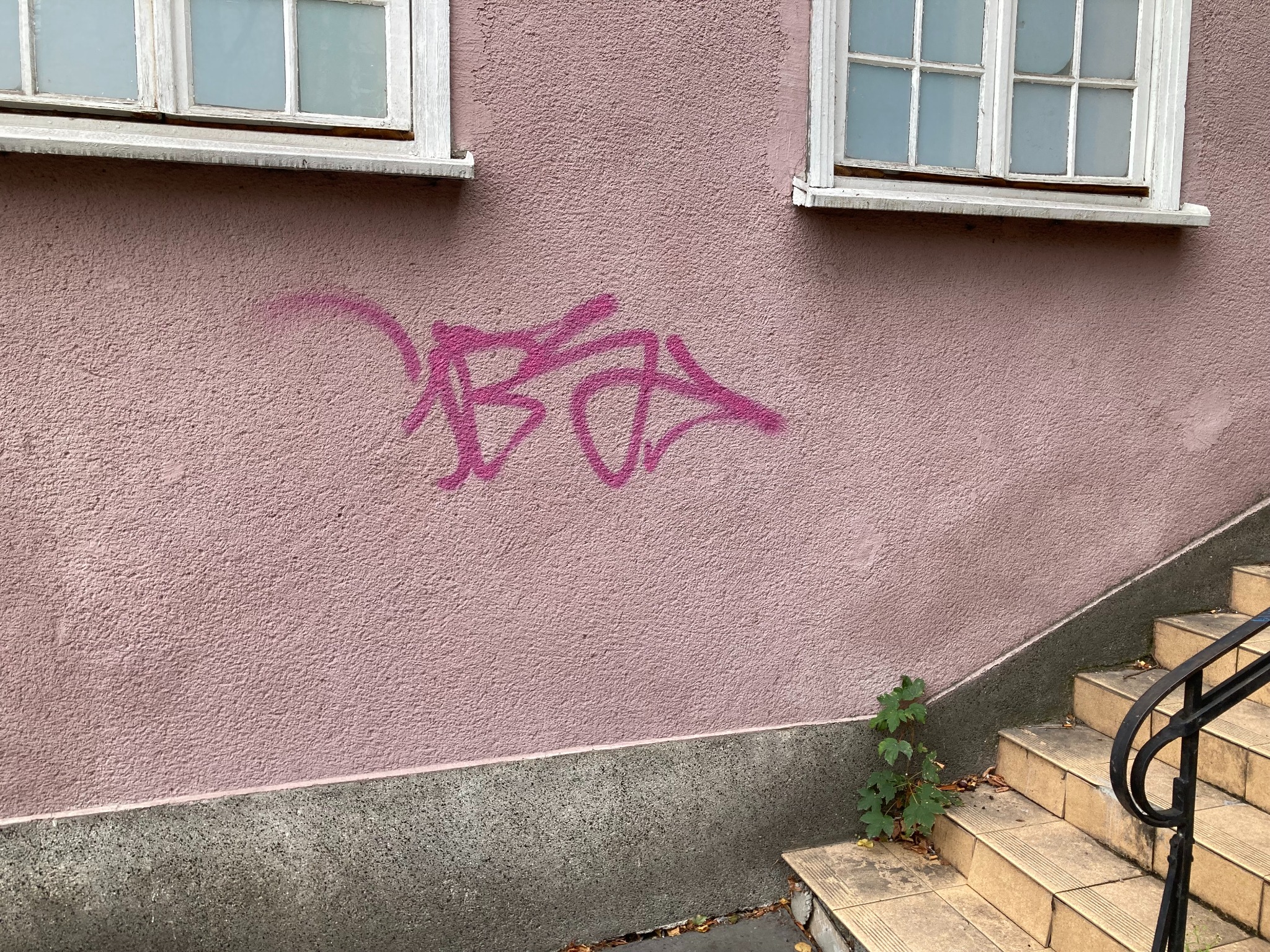 Różowa elewacja budynku z graffiti, widoczne okna z białymi ramami i schody z czarną barierką.