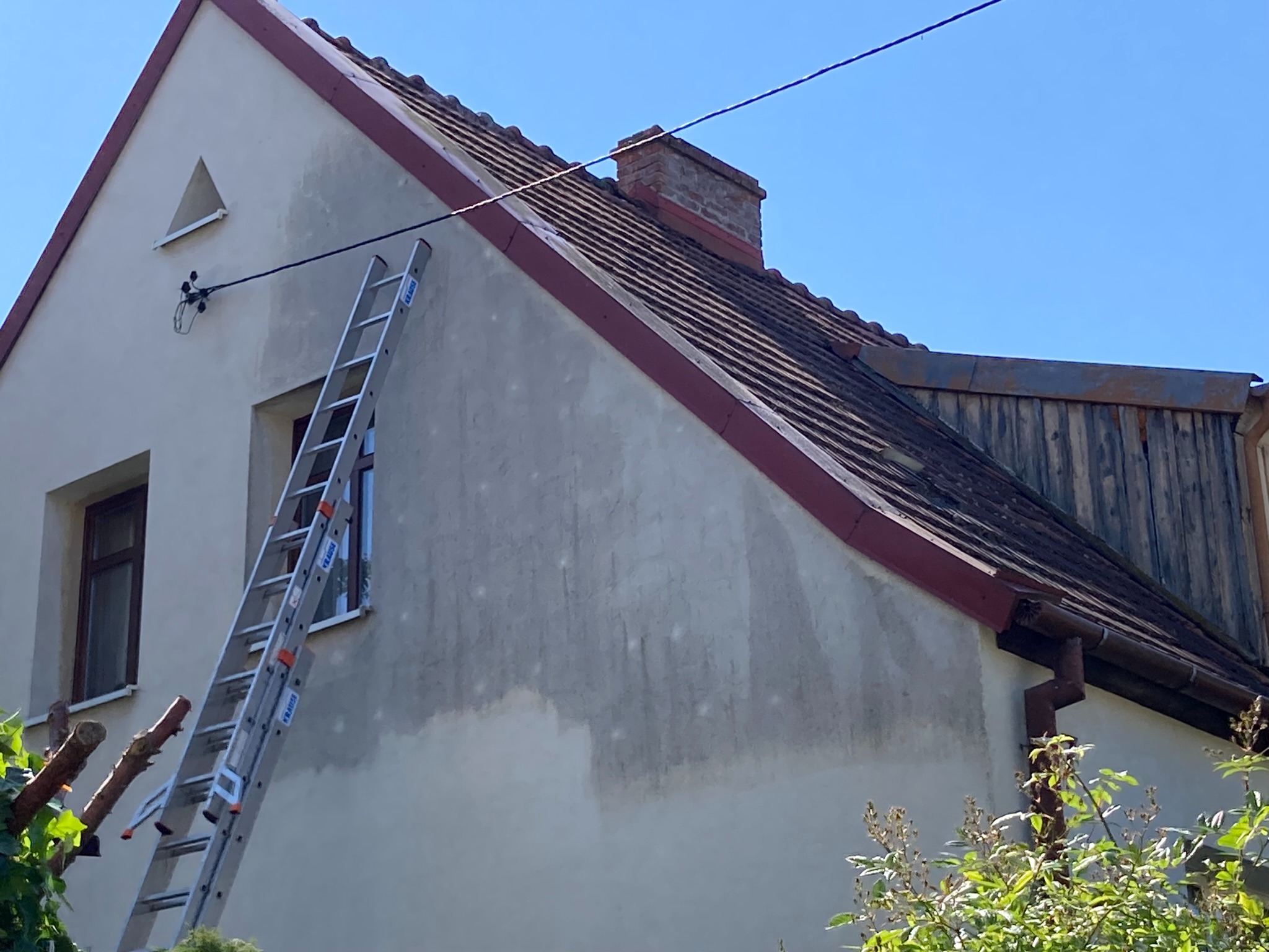 Ściana domu z widocznymi śladami po czyszczeniu, drabina oparta o ścianę, dach z czerwonymi obrzeżami, ceglany komin, błękitne niebo w tle.