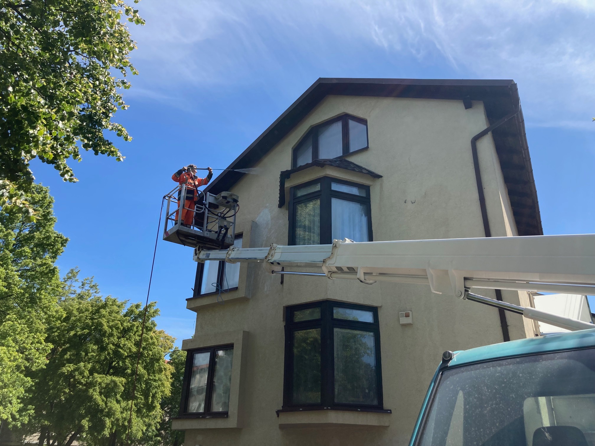 Pracownik w pomarańczowym kombinezonie na podnośniku koszowym czyści elewację dwupiętrowego domu strumieniem wody pod ciśnieniem, widoczny fragment nieumytego i umytego muru.