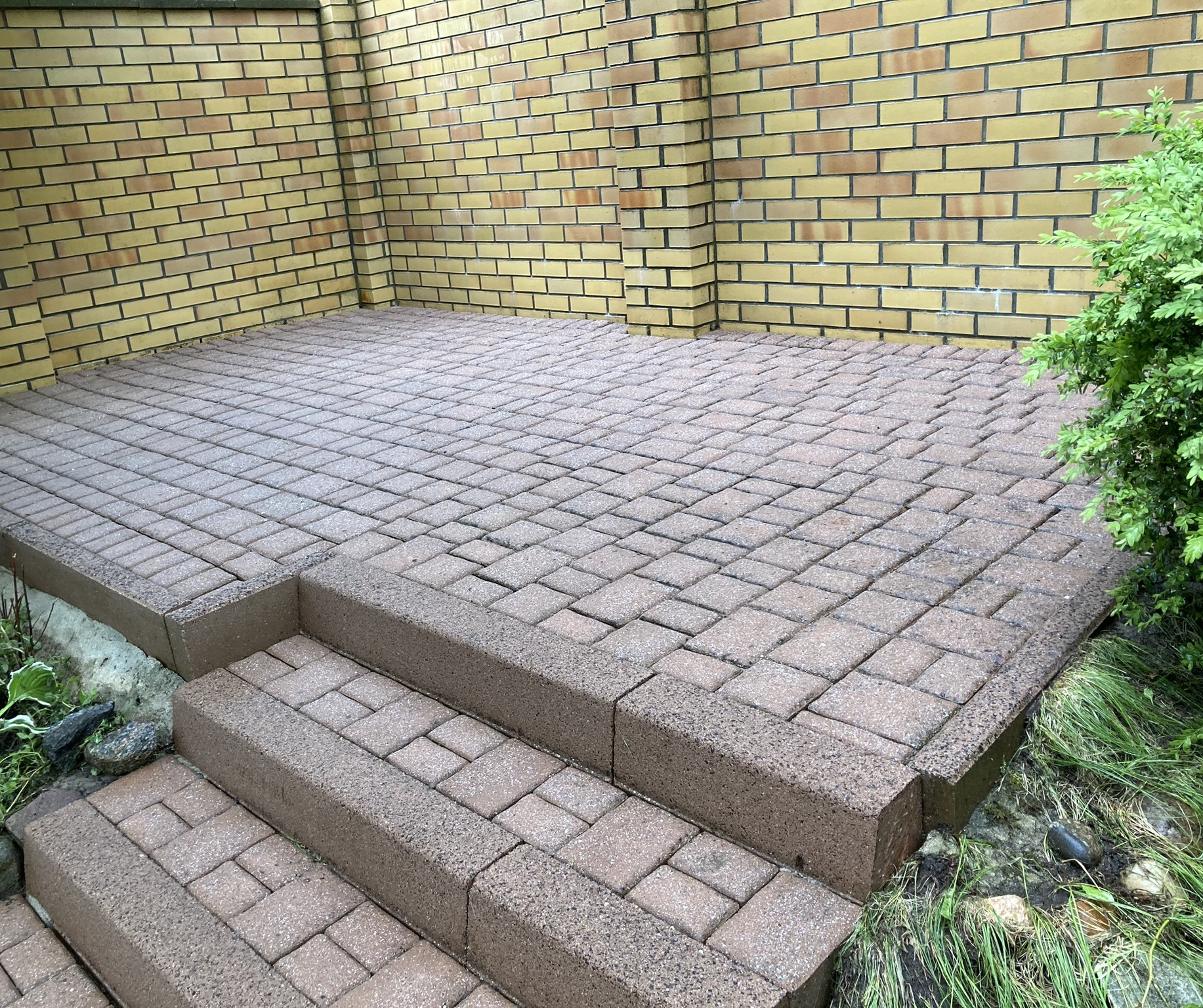 Brązowa kostka brukowa ułożona na schodach i podwyższonym tarasie, otoczona zielenią i żółtą ścianą z cegły.