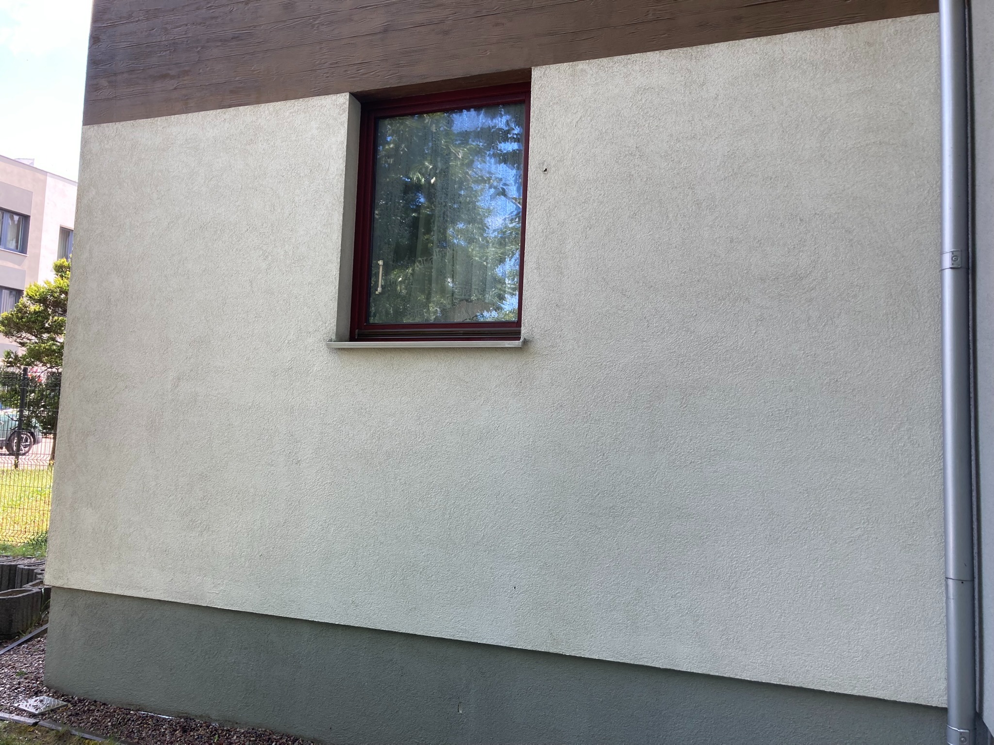 Fragment elewacji budynku z oknem w bordowej ramie i rynną, widoczna struktura tynku baranka i drewnopodobne wykończenie górnej części ściany.