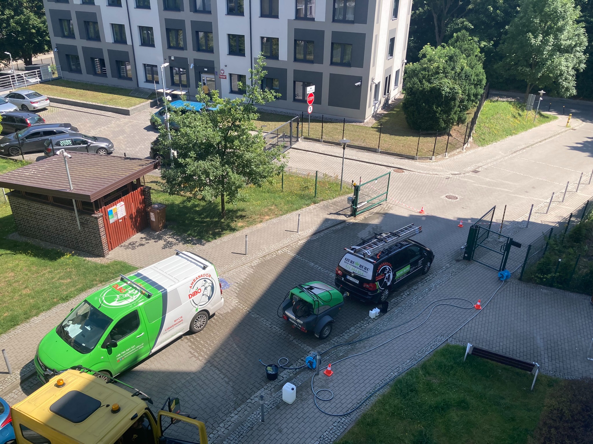 Widok z góry na zaplecze firmy sprzątającej na kostce brukowej przed budynkiem mieszkalnym: zielono-biały van, czarne auto z drabiną na dachu, przyczepa z urządzeniem ciśnieniowym, węże, wiadro...