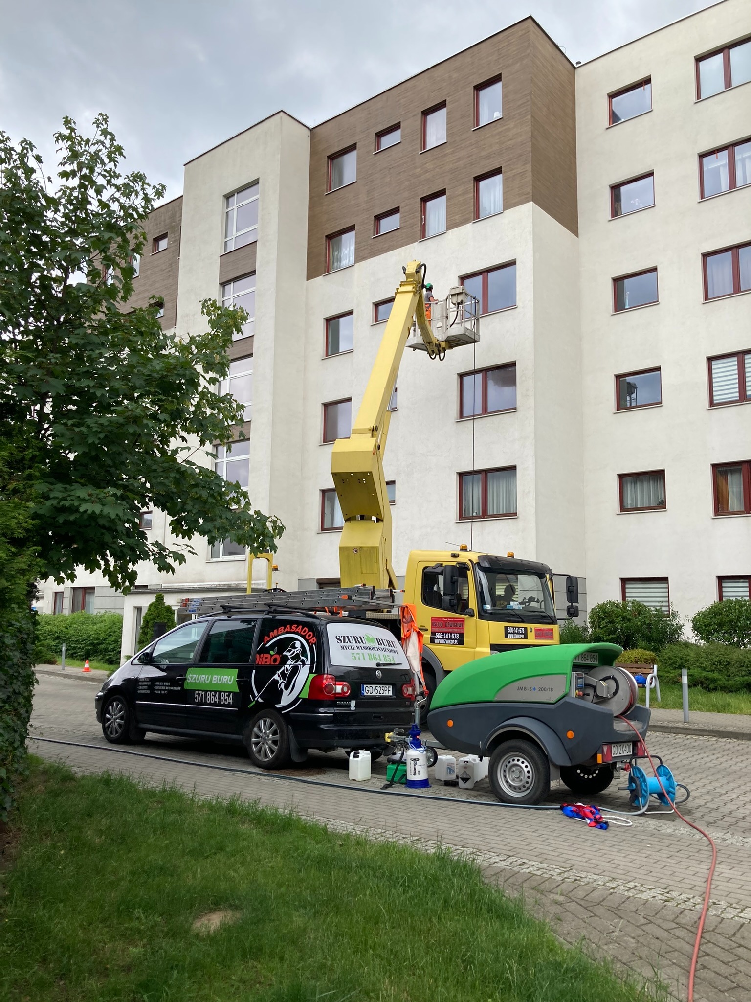 Pracownik na podnośniku koszowym czyści elewację budynku wielorodzinnego; widoczny samochód firmowy z logo 'Szuru Buru' i przyczepka ze sprzętem do czyszczenia.
