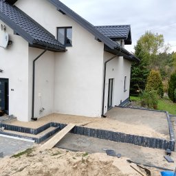 Norbert Nowakowski PaintHouse - Nowoczesny dom jednorodzinny z przygotowanym podłożem pod kostkę brukową na tarasie i chodniku, widoczny pies leżący przed wejściem.