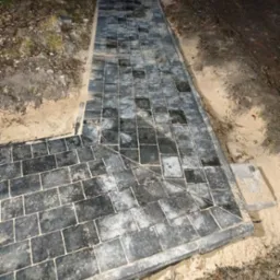 Ułożona ścieżka z kostki brukowej w odcieniach szarości, z widocznym piaskiem pomiędzy kostkami i na obrzeżach, świeżo po ułożeniu, z widocznym fragmentem trawnika po bokach.