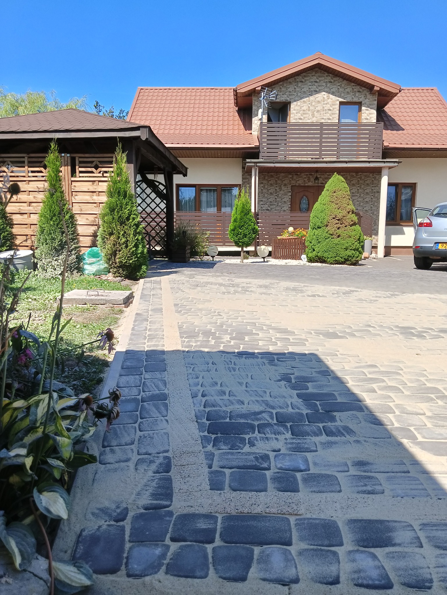 Nowo ułożona, szara kostka brukowa prowadząca do domu z brązowym dachem i drewnianą altaną po lewej stronie; widoczny piasek pomiędzy kostkami.