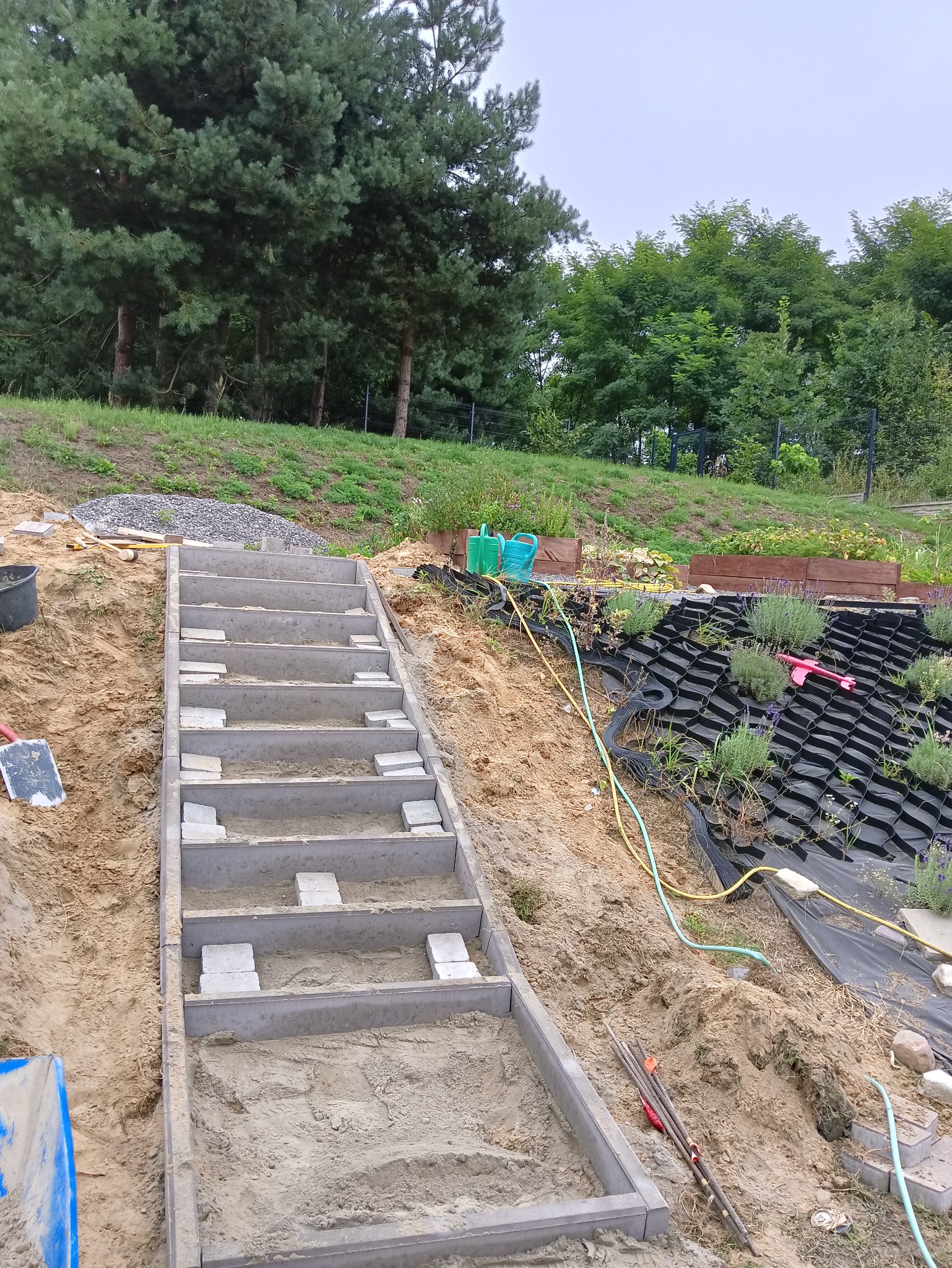 Betonowe schody ogrodowe w trakcie budowy na zboczu, widoczne szalunki, wypełnienie piaskiem i przygotowane stopnie z kostki, w tle drzewa i agrowłóknina na skarpie.