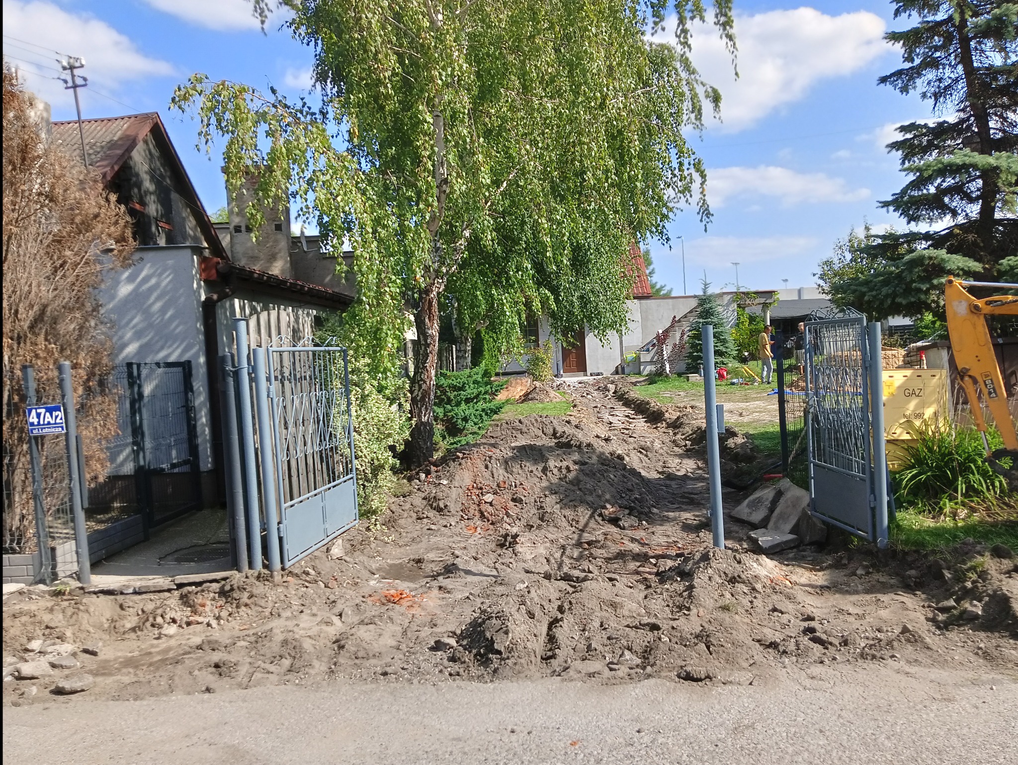 Przygotowanie podłoża pod kostkę brukową przed posesją z otwartą bramą i widocznym żółtym sprzętem budowlanym w tle.