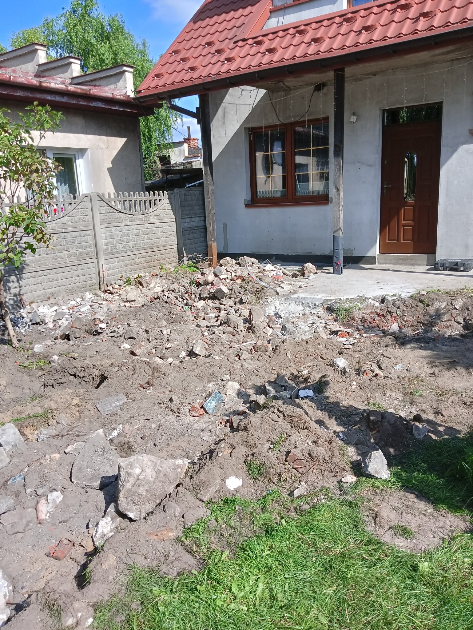 Przygotowanie podłoża pod kostkę brukową przed domem z czerwonym dachem, widoczne gruz, ziemia i fragment betonowego ogrodzenia.