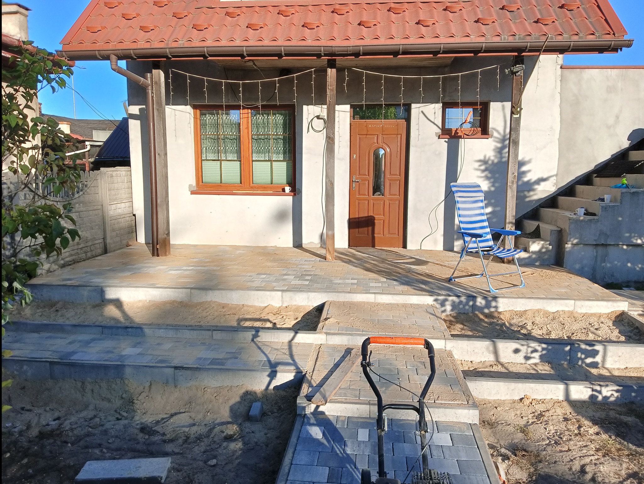 Nowo ułożona kostka brukowa przed domem z widocznym podjazdem, schodami i tarasem, częściowo jeszcze w piasku, z narzędziem do układania i krzesłem plażowym w tle.