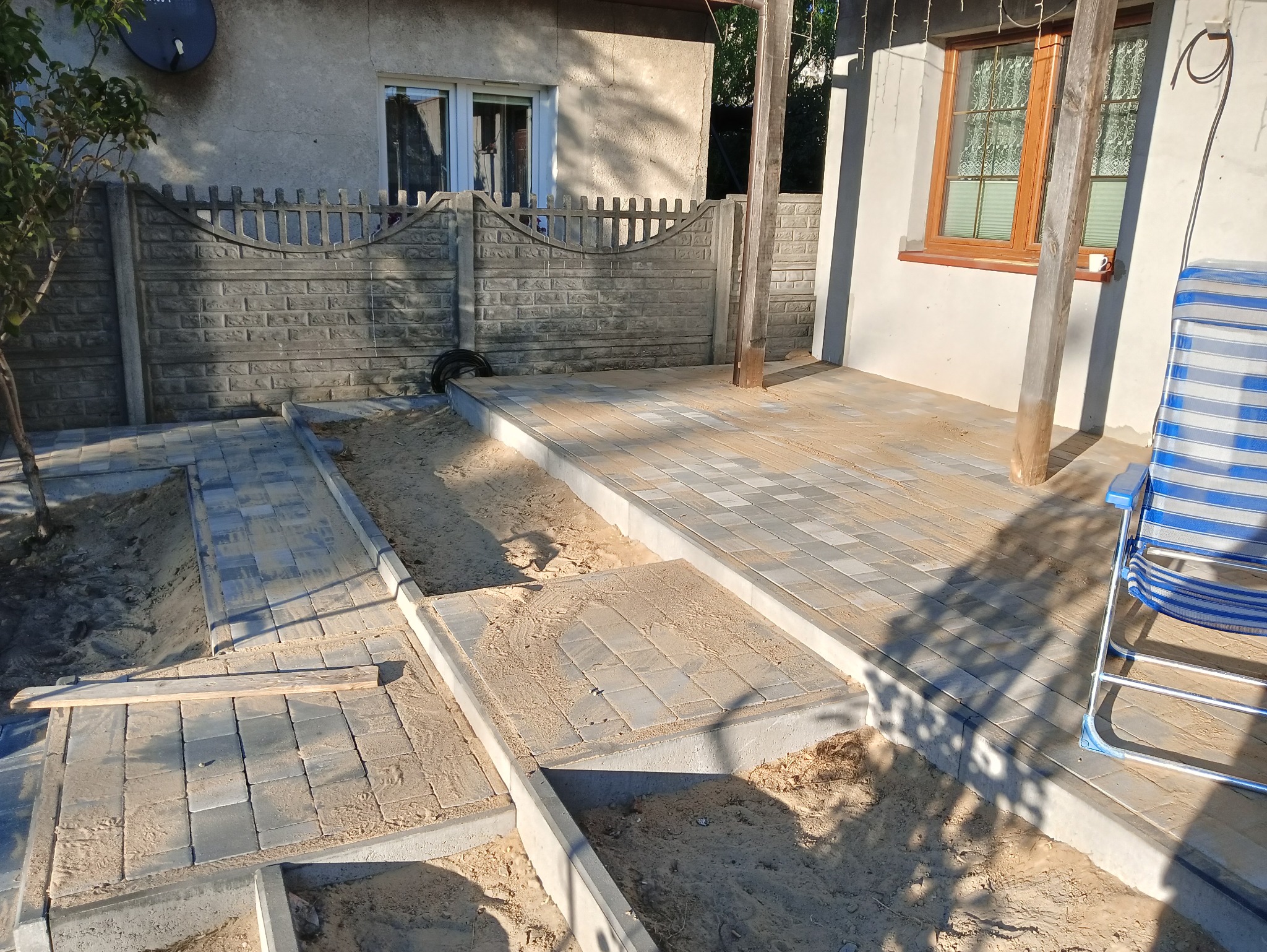 Fragment tarasu w trakcie budowy z szarych kostek brukowych, widoczne przygotowane podłoże z piasku, betonowe obrzeża i krzesło plażowe w tle.