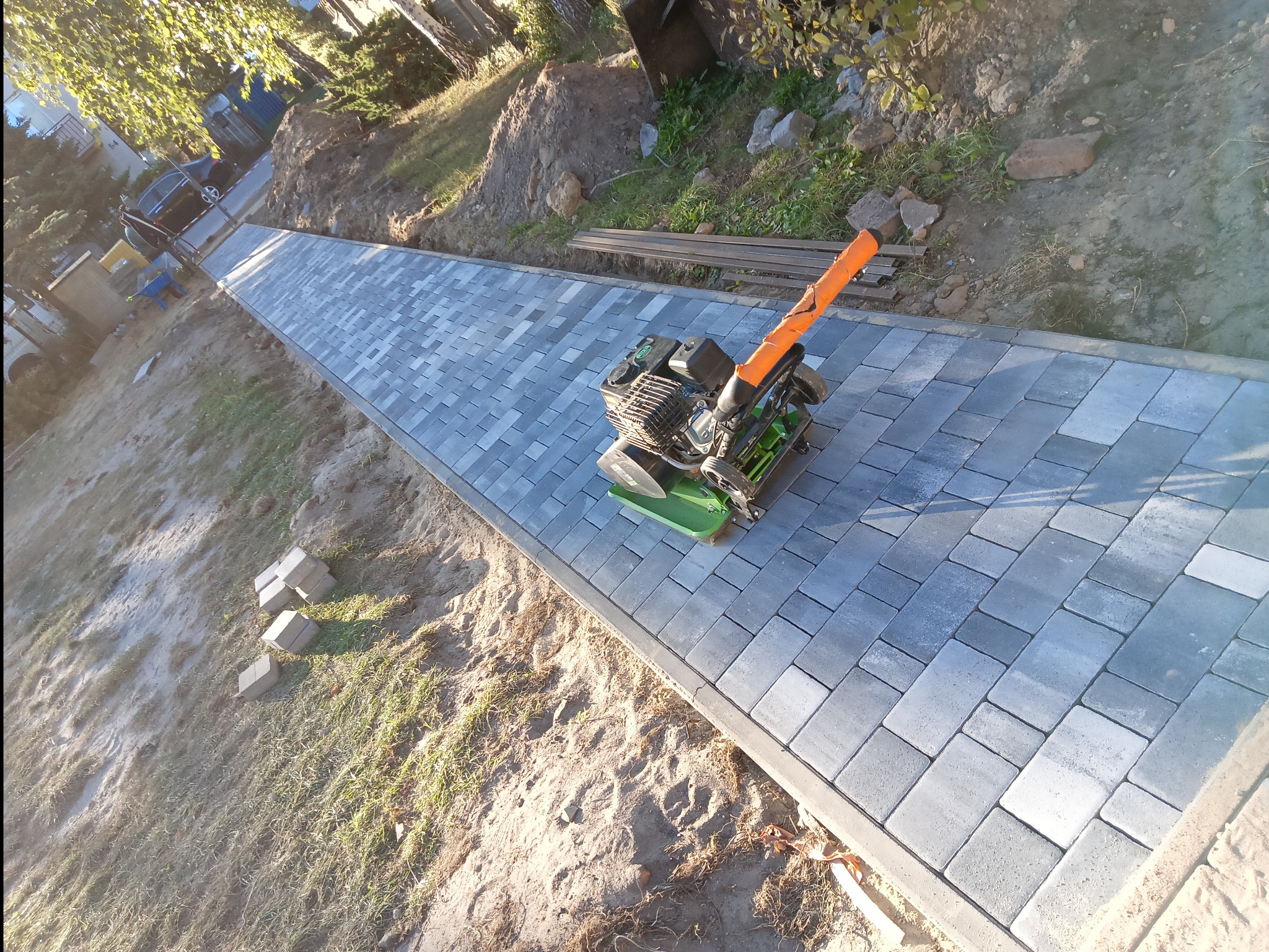 Świeżo ułożona, szara kostka brukowa na długim podjeździe, ubijana zagęszczarką; w tle widoczny stos kostki, trawa i częściowo wykopana ziemia.