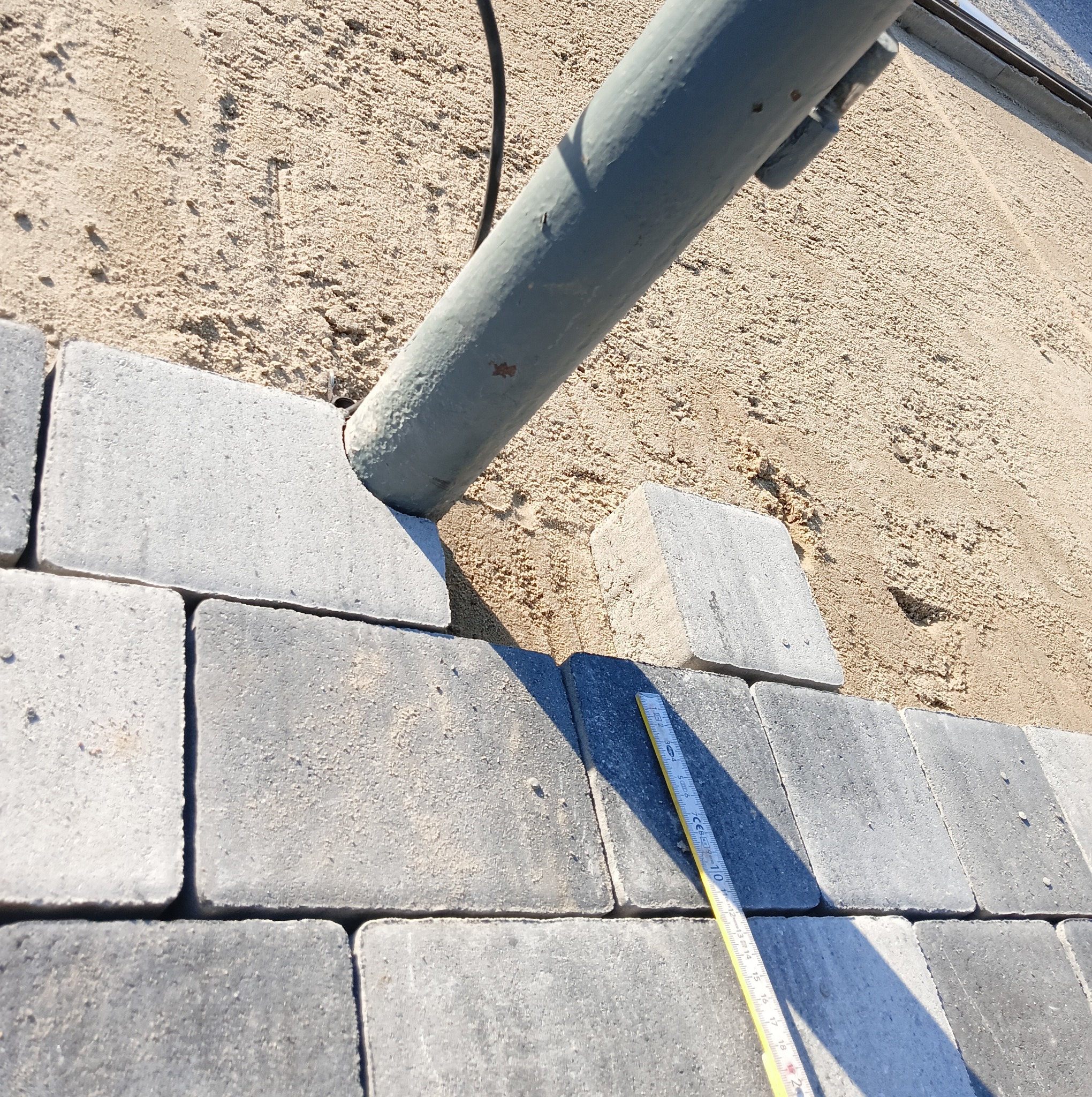 Szara kostka brukowa układana przy szarej rurze, z widocznym piaskiem w tle i żółtą miarką na kostce.