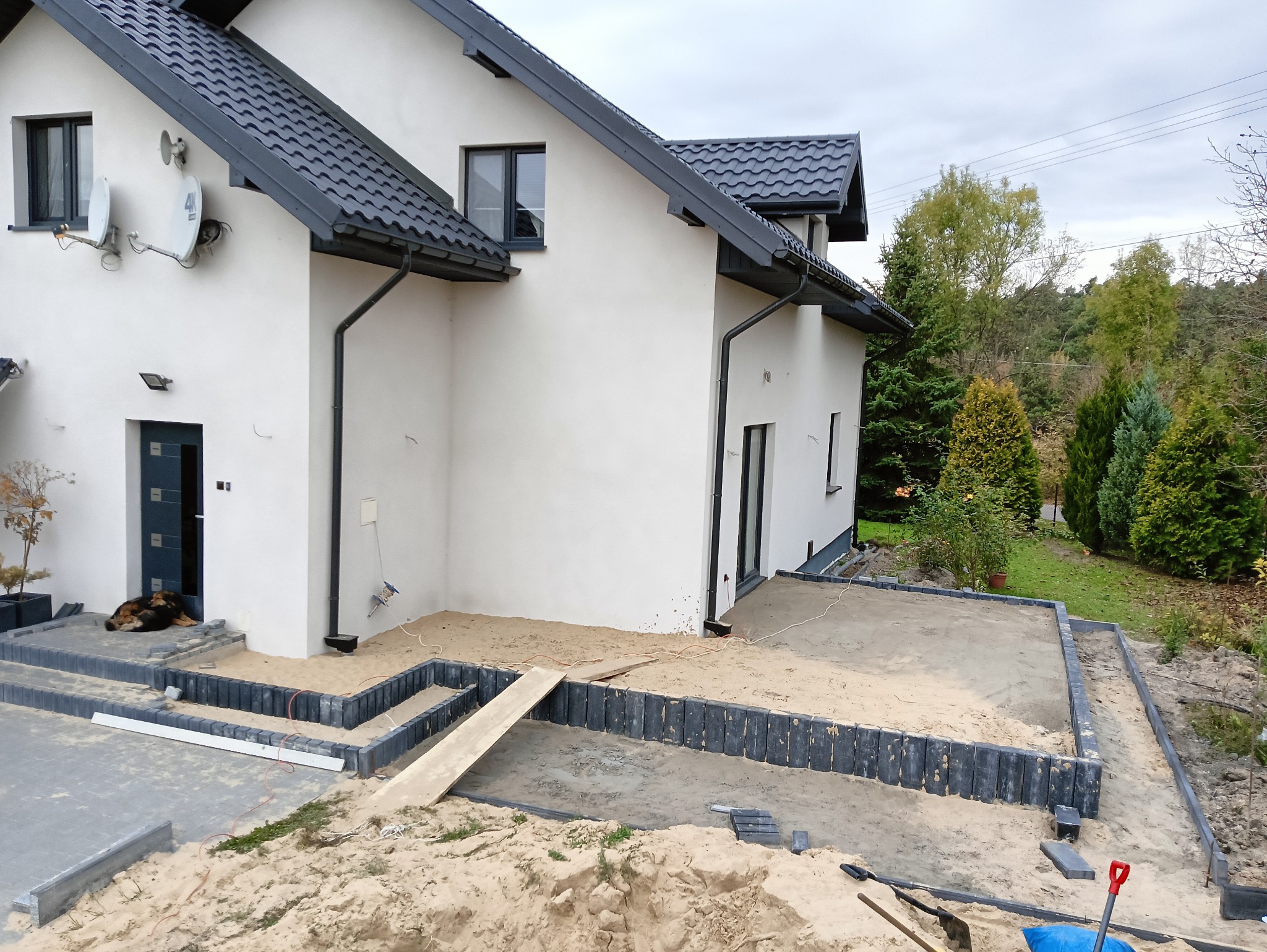 Nowoczesny dom jednorodzinny z przygotowanym podłożem pod kostkę brukową na tarasie i chodniku, widoczny pies leżący przed wejściem.