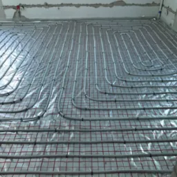 Rozłożona instalacja ogrzewania podłogowego na folii izolacyjnej z czerwoną siatką w surowym wnętrzu budynku w trakcie budowy.