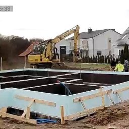 SOLID HAUS ADAM KOSATER - Fundamenty domu w trakcie budowy, z widoczną izolacją termiczną i zbrojeniami. W tle pracuje koparka, a obok fundamentów stoją pracownicy budowlani.