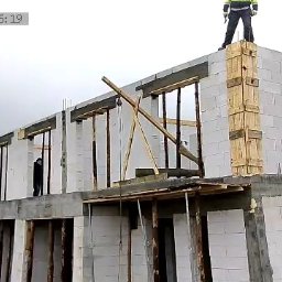 SOLID HAUS ADAM KOSATER - Wznoszenie dwukondygnacyjnego domu z białych bloczków, widoczne drewniane szalunki okienne i słupów. Na dachu stoi osoba w odblaskowej kamizelce.