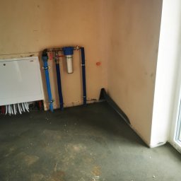 SOLID HAUS ADAM KOSATER - Nowa instalacja wodna z filtrem i rozdzielnicą w surowym wnętrzu, widoczne rury w kolorze niebieskim i białe przewody, świeżo wylana posadzka.