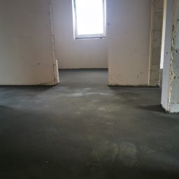 SOLID HAUS ADAM KOSATER - Wylana, ciemna posadzka w surowym wnętrzu z oknem. Widoczne fragmenty ścian i słupów. Na pierwszym planie elementy szalunku.