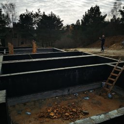 SOLID HAUS ADAM KOSATER - Fundamenty domu jednorodzinnego z betonu komórkowego, widok z góry, na tle lasu i pochmurnego nieba. Na górze kadru mężczyzna robiący zdjęcie telefonem.