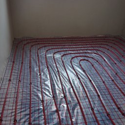 SOLID HAUS ADAM KOSATER - Czerwone rury ogrzewania podłogowego ułożone na srebrnej folii z siatką, przymocowane czarnymi klipsami, widok z góry w rogu pomieszczenia.