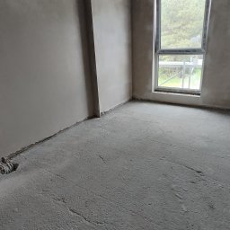 SOLID HAUS ADAM KOSATER - Surowe wnętrze pokoju z oknem, ściany pokryte gładzią, betonowa podłoga z widocznym otworem na instalacje. W tle widoczne rusztowanie za oknem.