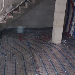SOLID HAUS ADAM KOSATER - Instalacja ogrzewania podłogowego z rurami na folii z siatką, widok z góry, surowy stan, betonowe schody w tle, kolumna z bloczków.