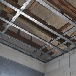 SOLID HAUS ADAM KOSATER - Sufit podwieszany w trakcie montażu, widoczne profile metalowe, izolacja z wełny mineralnej i fragment ściany z białych bloczków. Ujęcie z dołu, surowy stan.