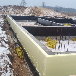 SOLID HAUS ADAM KOSATER - Fundamenty domu w trakcie budowy, ocieplone styropianem, widoczne zbrojenia i folia izolacyjna. W tle ziemia i drzewa.