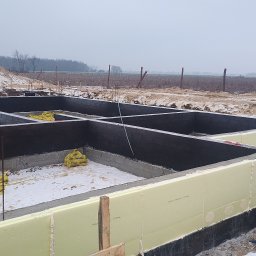 SOLID HAUS ADAM KOSATER - Fundamenty domu jednorodzinnego z izolacją termiczną, widoczne zbrojenia i folia. Zimowy krajobraz w tle, pochmurne niebo. Etap budowy przed wylaniem płyty fundamentowej.