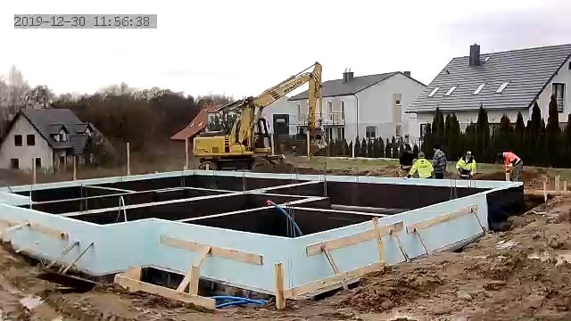 Fundamenty domu w trakcie budowy, z widoczną izolacją termiczną i zbrojeniami. W tle pracuje koparka, a obok fundamentów stoją pracownicy budowlani.