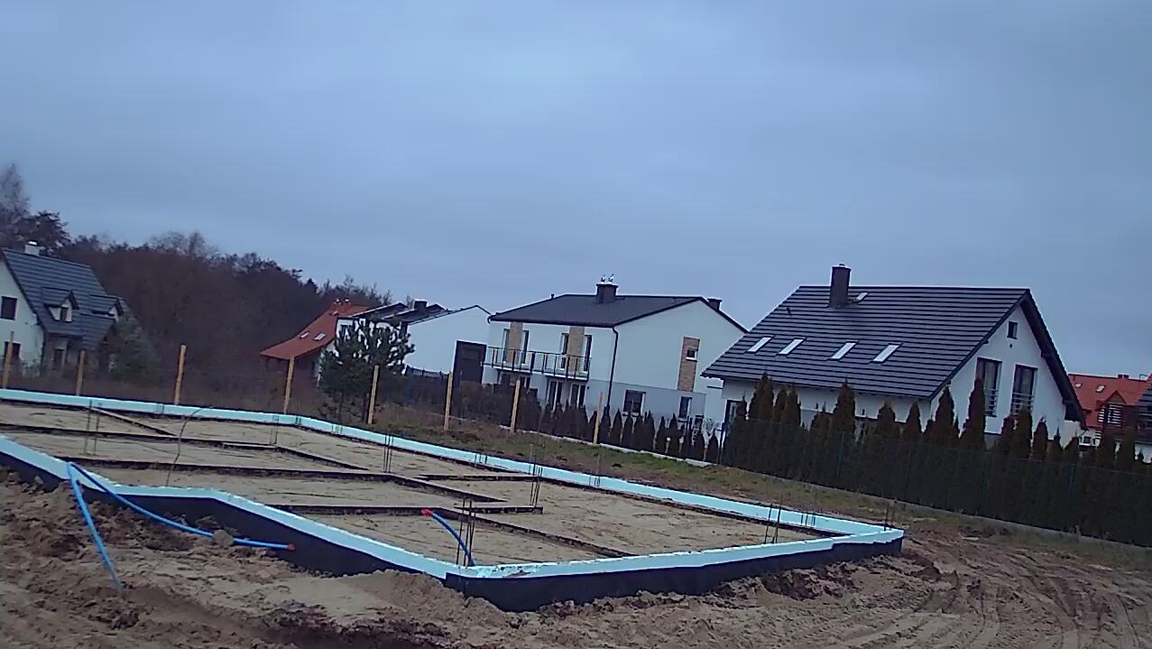Fundamenty domu jednorodzinnego w trakcie budowy, z widoczną izolacją termiczną i przygotowaniem pod instalacje. W tle zabudowa mieszkalna i pochmurne niebo.