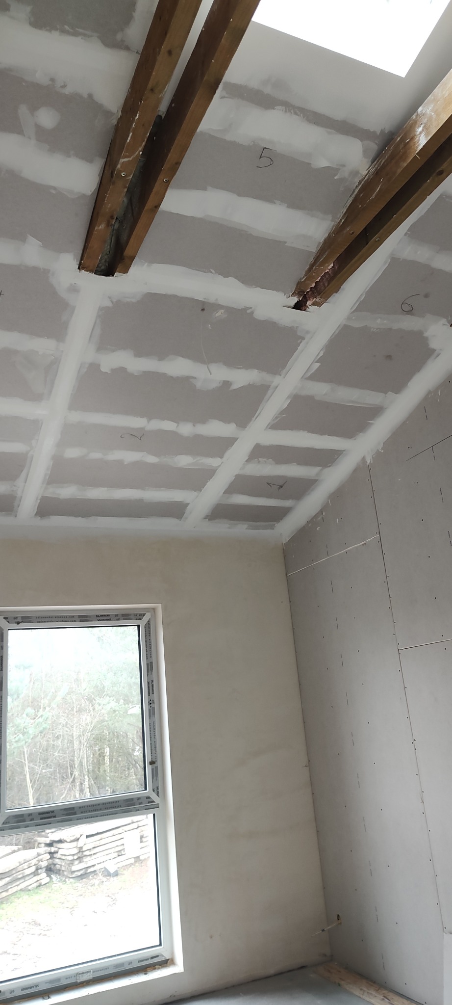 Wnętrze w trakcie prac wykończeniowych: sufit z płyt gipsowo-kartonowych z widocznymi drewnianymi belkami, ściany przygotowane pod gładzie, okno z widokiem na zewnątrz.
