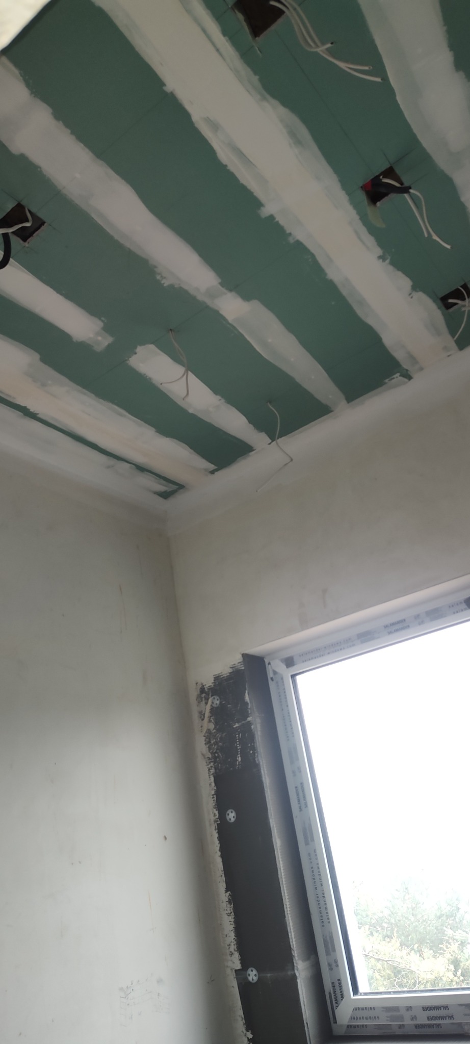 Sufit z płyt gipsowo-kartonowych, częściowo zaszpachlowany, z widocznymi przewodami elektrycznymi. Ściana z oknem w tle. Widoczne niedoskonałości wykończenia.