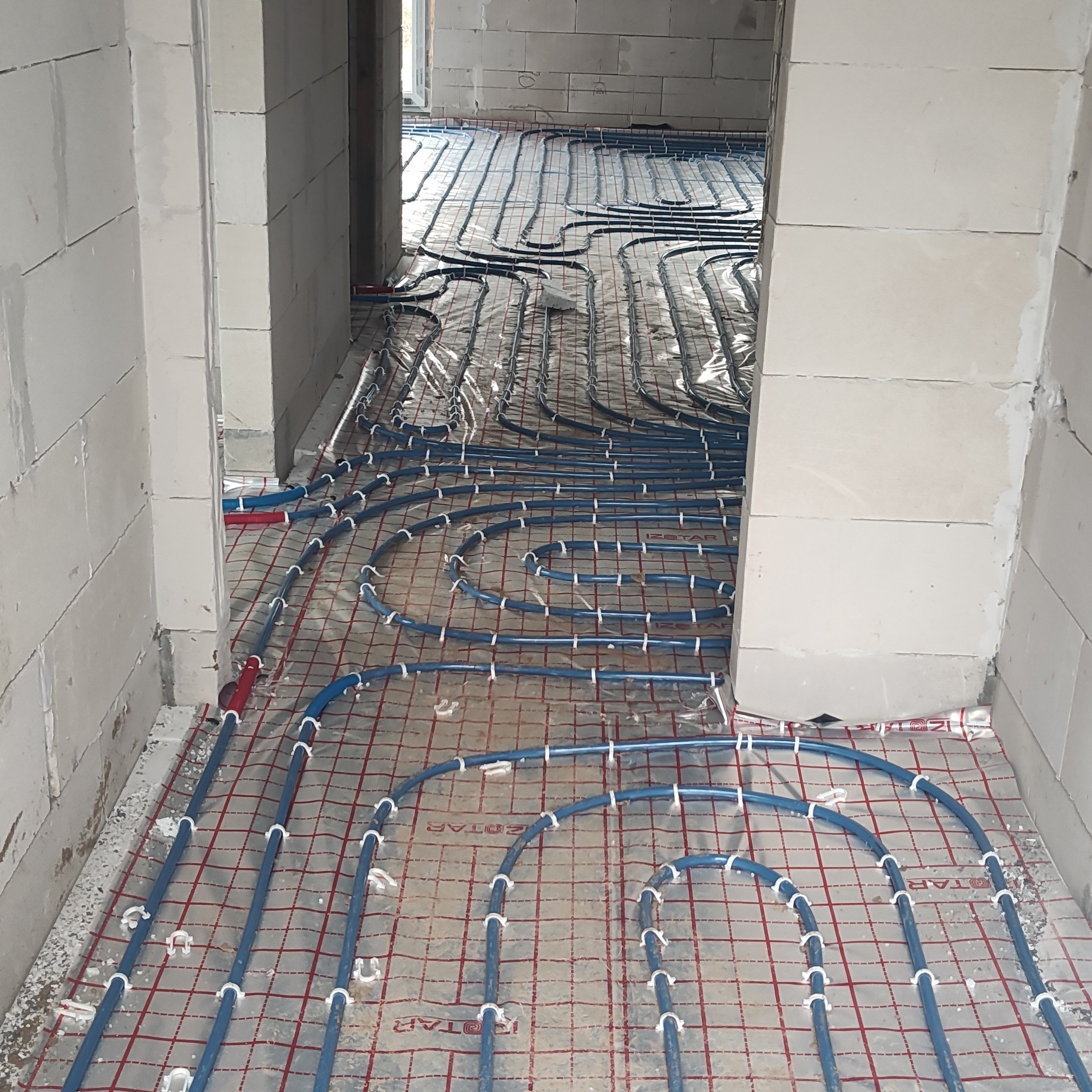 Niebieskie rury ogrzewania podłogowego, zamocowane na folii z czerwoną siatką, w surowym wnętrzu z białymi ścianami z betonu komórkowego.