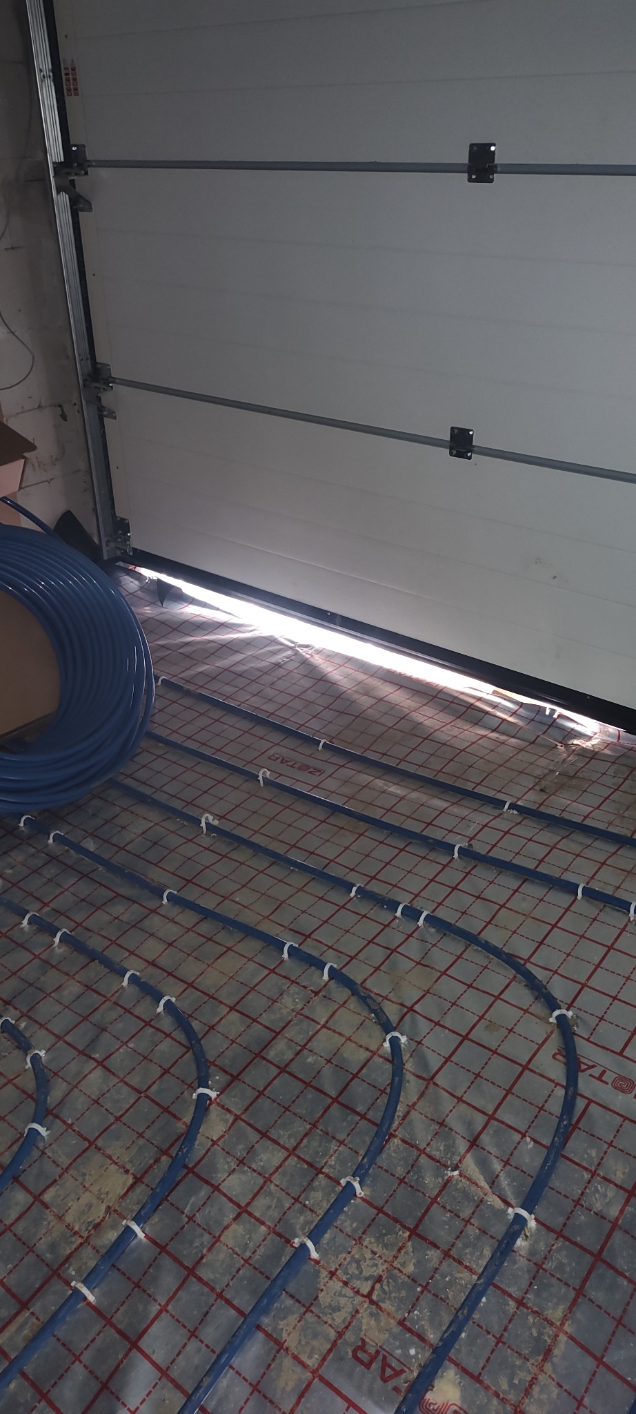 Niebieskie rury ogrzewania podłogowego, przymocowane białymi uchwytami do folii z czerwoną siatką, widoczne przy otwartych drzwiach garażowych.