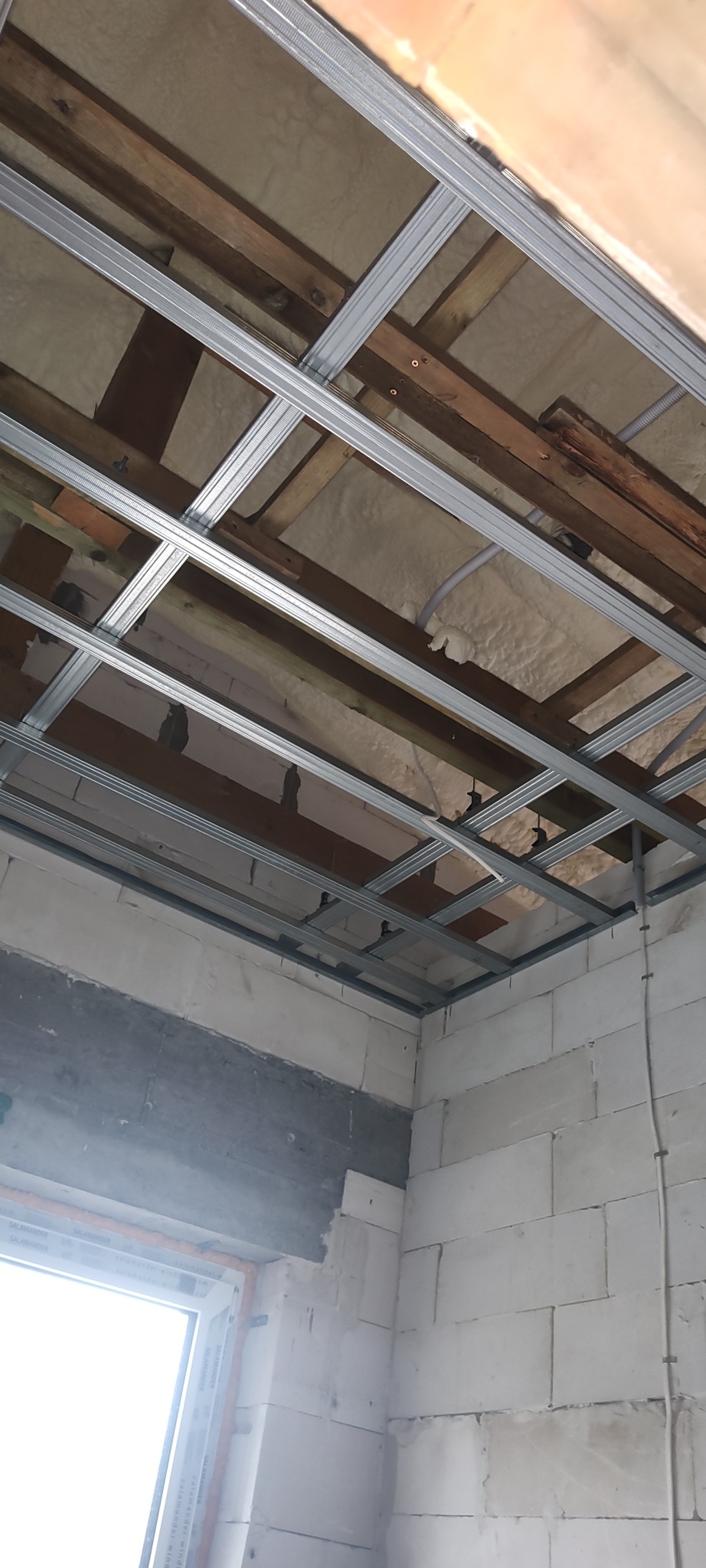 Sufit podwieszany w trakcie montażu, widoczne profile metalowe, izolacja z wełny mineralnej i fragment ściany z białych bloczków. Ujęcie z dołu, surowy stan.