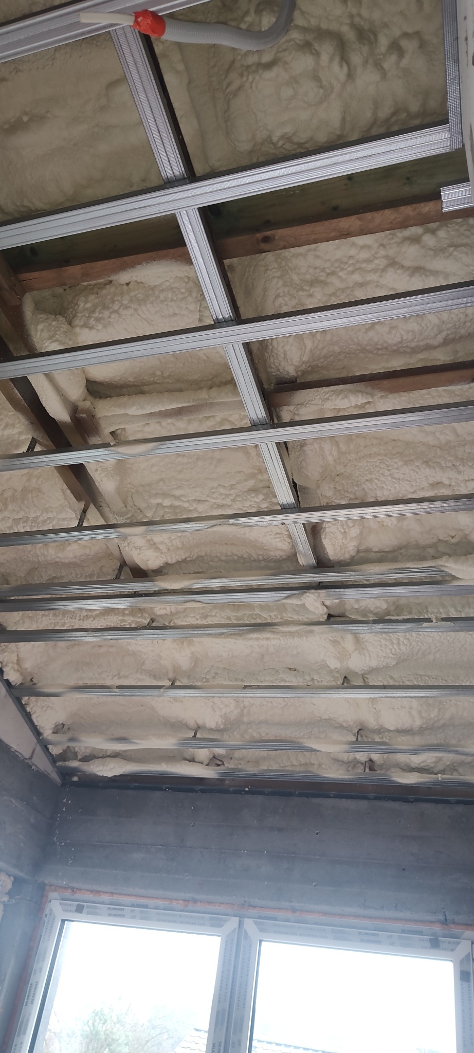 Poddasze z widoczną pianą izolacyjną między krokwiami i metalową konstrukcją sufitu podwieszanego, okno w dolnej części kadru.