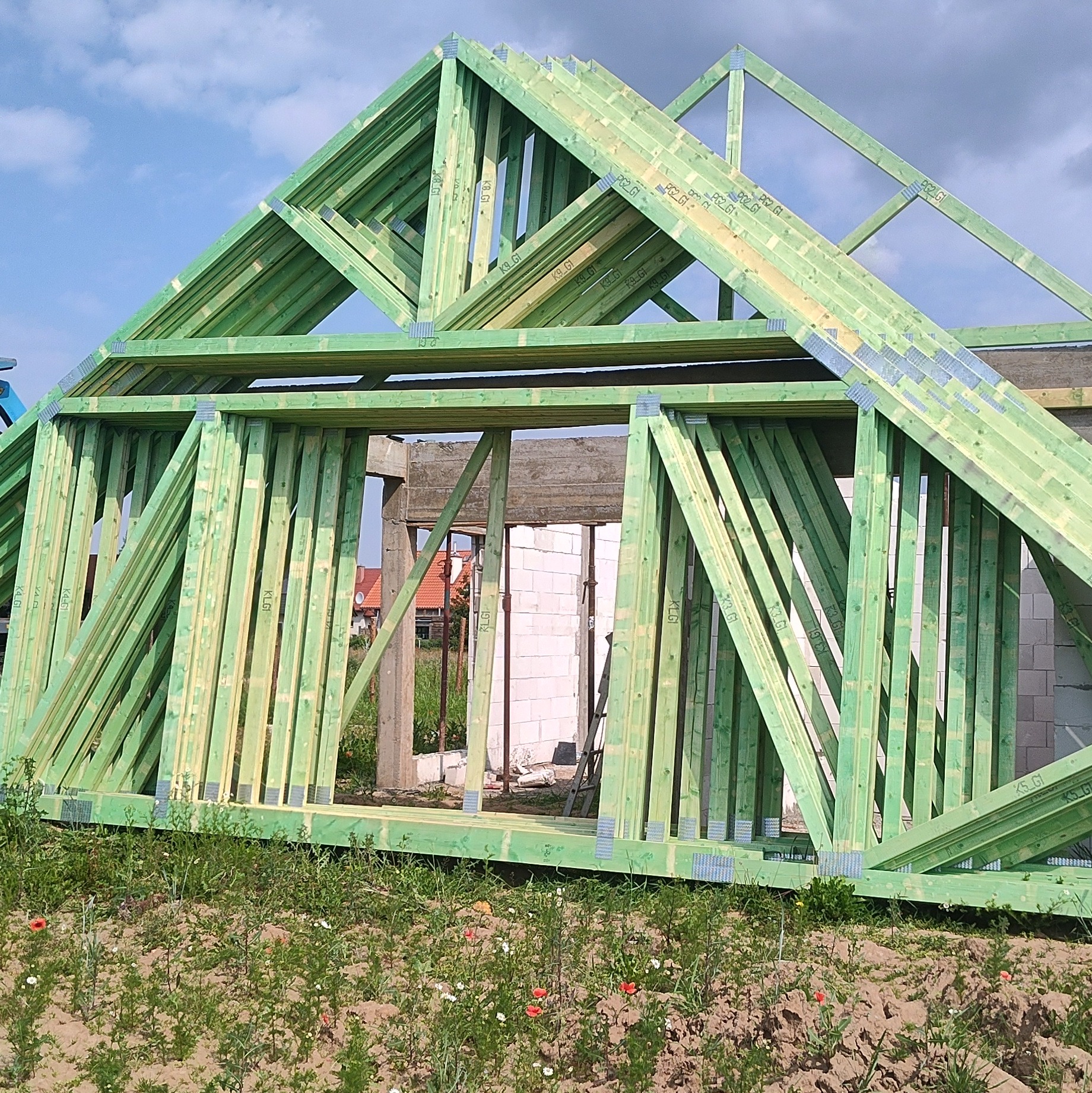 Konstrukcja dachu z impregnowanego drewna, widoczna na tle częściowo wymurowanego budynku i błękitnego nieba z chmurami. Elementy połączone metalowymi płytkami.