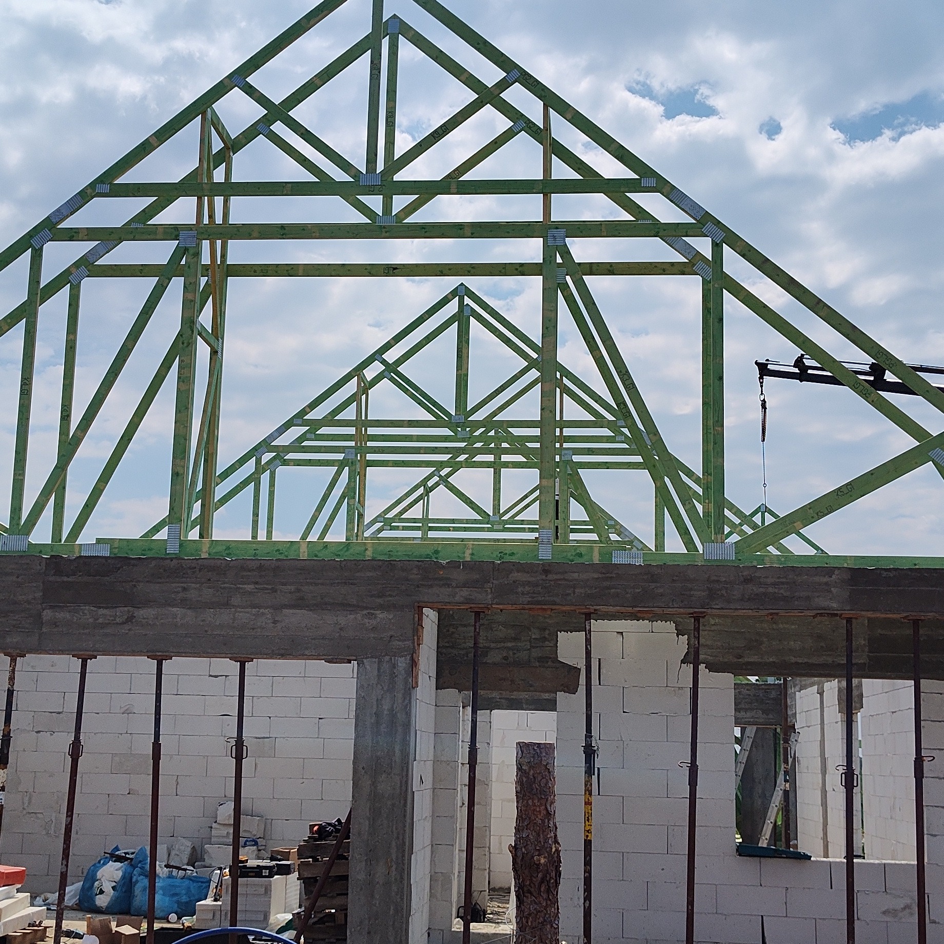 Konstrukcja dachu z zielonego drewna na domu w trakcie budowy, widok z dołu na tle błękitnego nieba z chmurami. Widoczne białe bloczki i podpory.