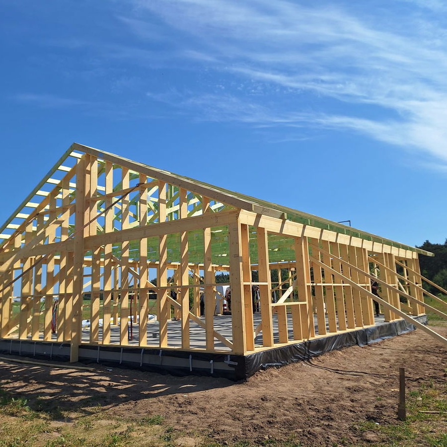 Surowa konstrukcja domu szkieletowego w słoneczny dzień. Widoczne drewniane elementy i fundamenty. Budowa w toku, na tle błękitnego nieba i zieleni.