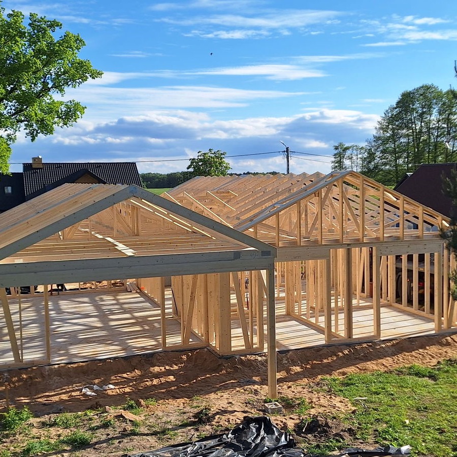 Konstrukcja szkieletowa domu w trakcie budowy, widoczne drewniane elementy ścian i dachu na tle zieleni i błękitnego nieba. Etap surowy otwarty.