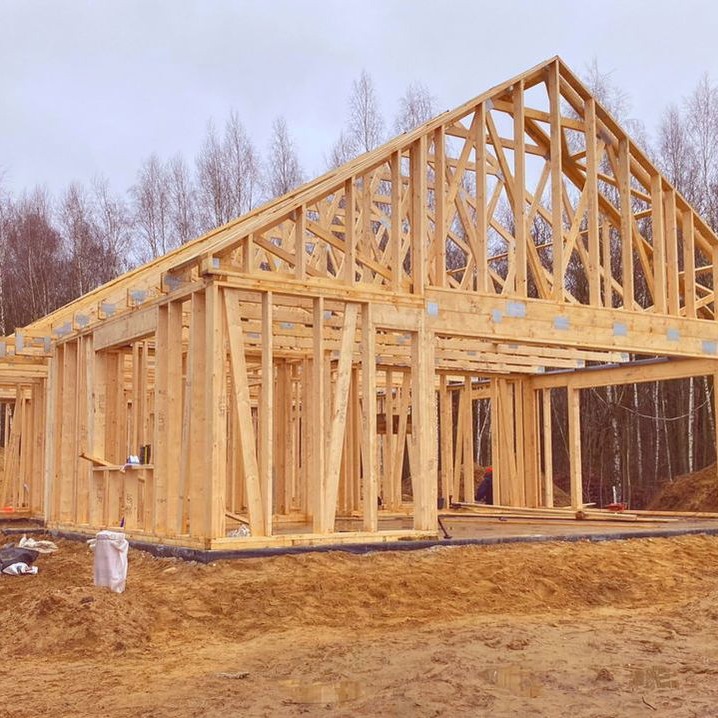 Surowa konstrukcja drewniana domu w trakcie budowy, widoczne elementy szkieletu i więźby dachowej na tle drzew i pochmurnego nieba. Etap wznoszenia ścian.