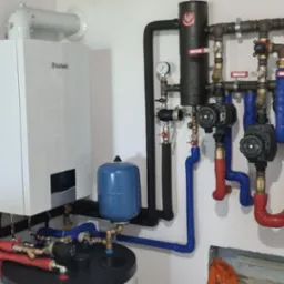 Nowoczesny, biały piec gazowy marki Vaillant z podłączonymi rurami w kolorze niebieskim i czerwonym, obok widoczny czarny filtr z manometrem oraz szara ściana z gniazdkiem elektrycznym.