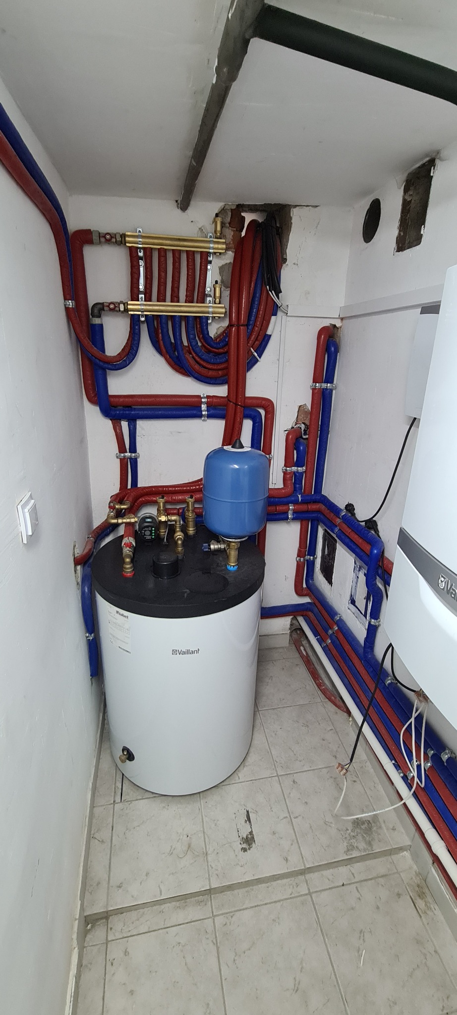 Bojler Vaillant z niebieskim zbiornikiem wyrównawczym, podłączony do instalacji z rurami w izolacji czerwonej i niebieskiej, zamontowany w małym pomieszczeniu z płytkami na podłodze.