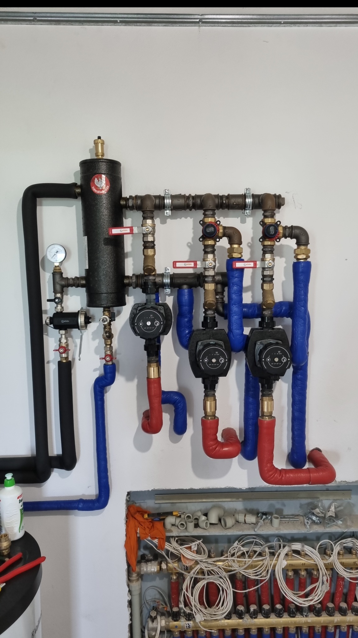 Rozbudowana instalacja centralnego ogrzewania z kotłem, pompami obiegowymi i rurami w izolacji czerwonej i niebieskiej, widoczne zawory i manometr, dolna część instalacji w trakcie montażu...