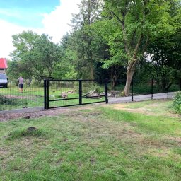 PanelExpress Ogrodzenia - Czarna brama panelowa z siatki na betonowej podmurówce, widok na posesję z zielonym trawnikiem, drzewami i zaparkowanym białym dostawczakiem w tle, podczas montażu przez pracownika.