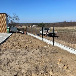 PanelExpress Ogrodzenia - Montaż ogrodzenia panelowego na betonowej podmurówce. Pracownik przy ciężarówce. Widok na otwarty teren i budynek z drewnianymi elementami.